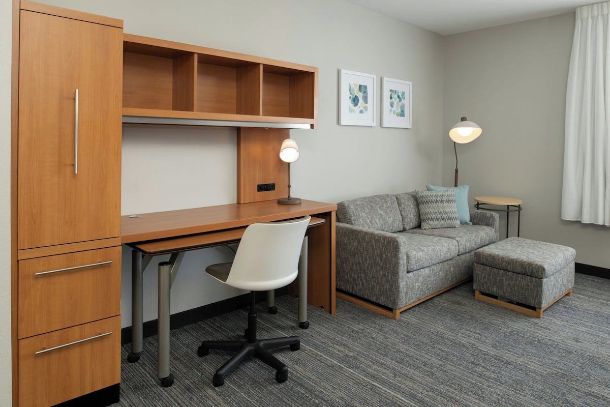 A compact hotel room with a wooden desk, a white office chair, a grey sofa, and a small ottoman. There are two wall-mounted lamps and two framed pictures on the wall.