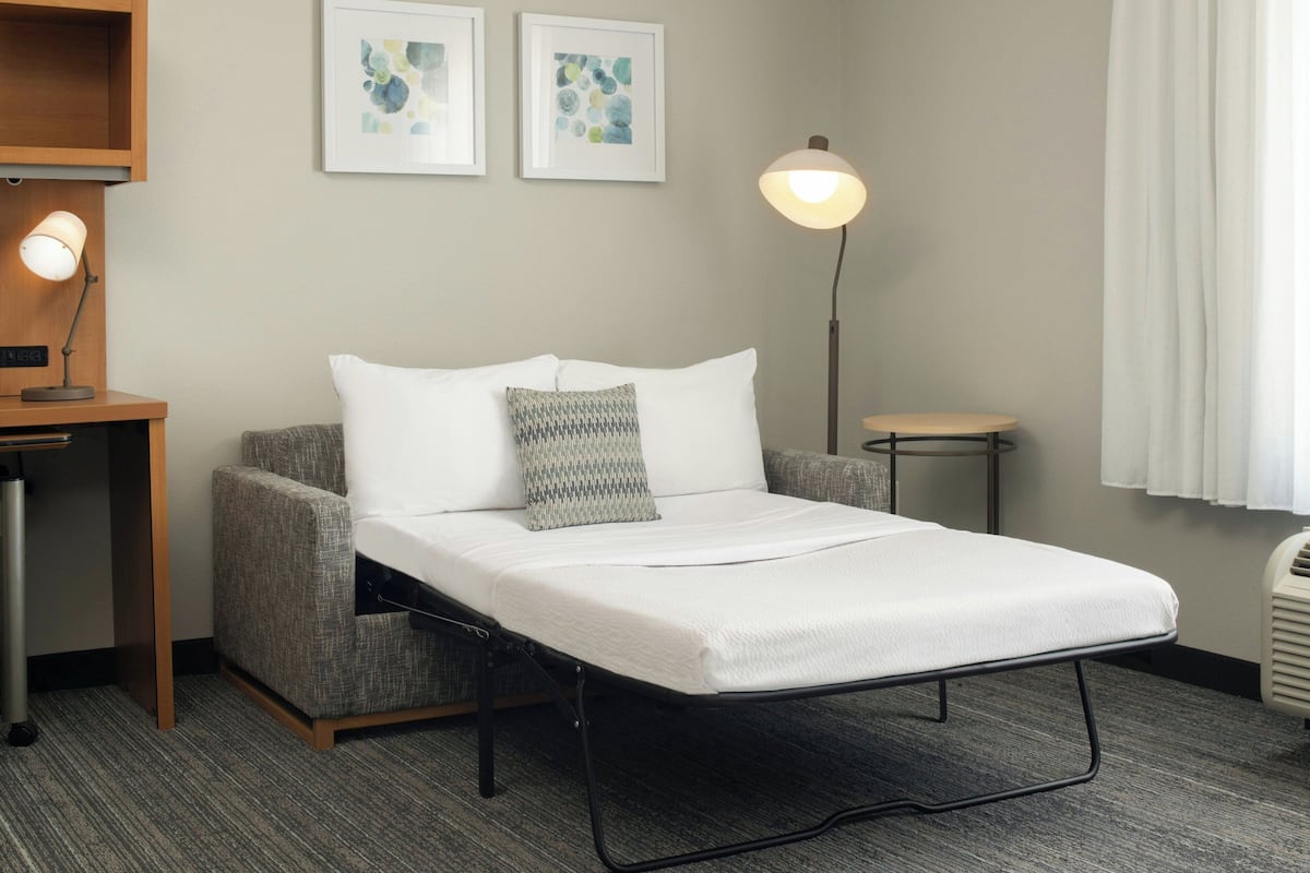 A single bed with a metal frame, a grey upholstered armchair, a small bedside table with a lamp, and two framed abstract artworks on the wall.
