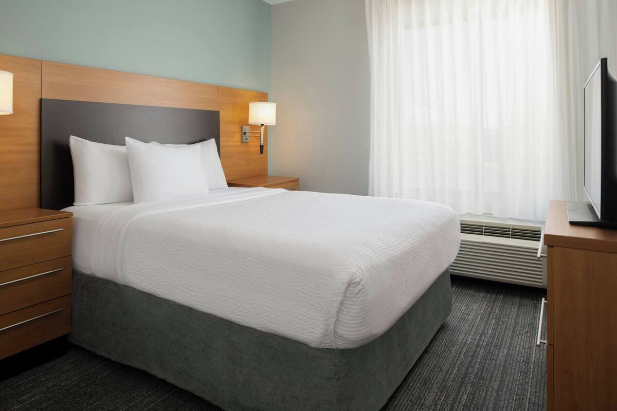 A hotel room with a large bed, a wooden headboard, a nightstand with a lamp, a television on a wooden cabinet, and a window with sheer curtains.