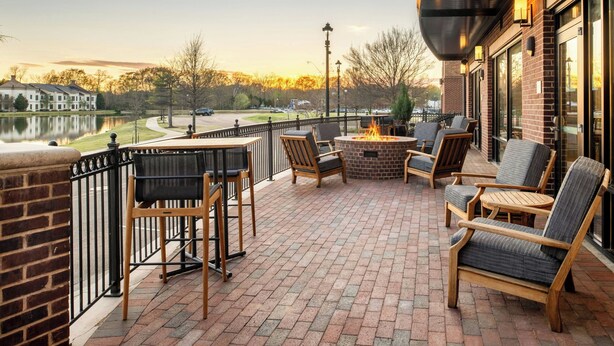 An outdoor patio with brick flooring, wooden chairs, a fire pit, and a view of a lake and buildings in the background. - TownePlace Suites Jackson Ridgeland/Township at Colony Park (Ridgeland)
