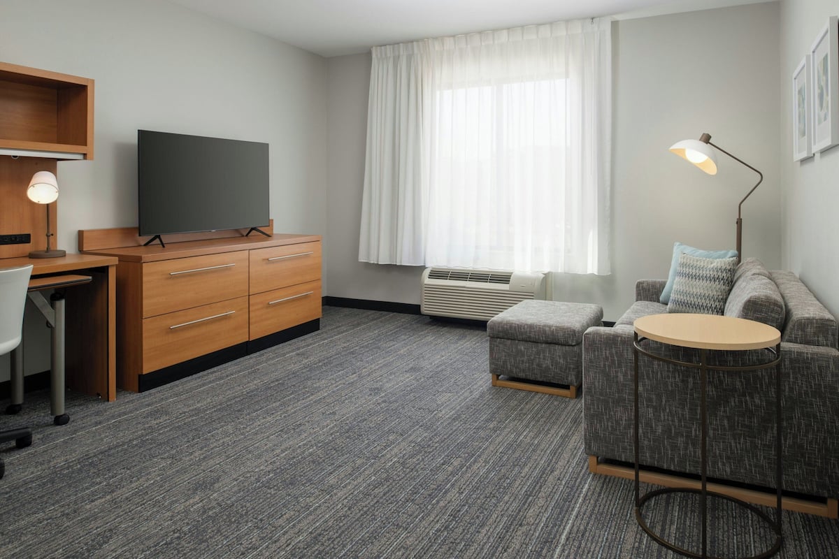 A modern hotel room with a wooden desk, a flat-screen TV, a grey sofa, a round side table, and a large window with sheer curtains.