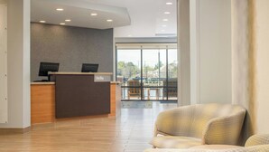 A reception area with a wooden reception desk, two monitors, and a "Hello" sign. There is a seating area with patterned chairs and a view of the outdoors through large windows. - TownePlace Suites Jackson Ridgeland/Township at Colony Park (Ridgeland)