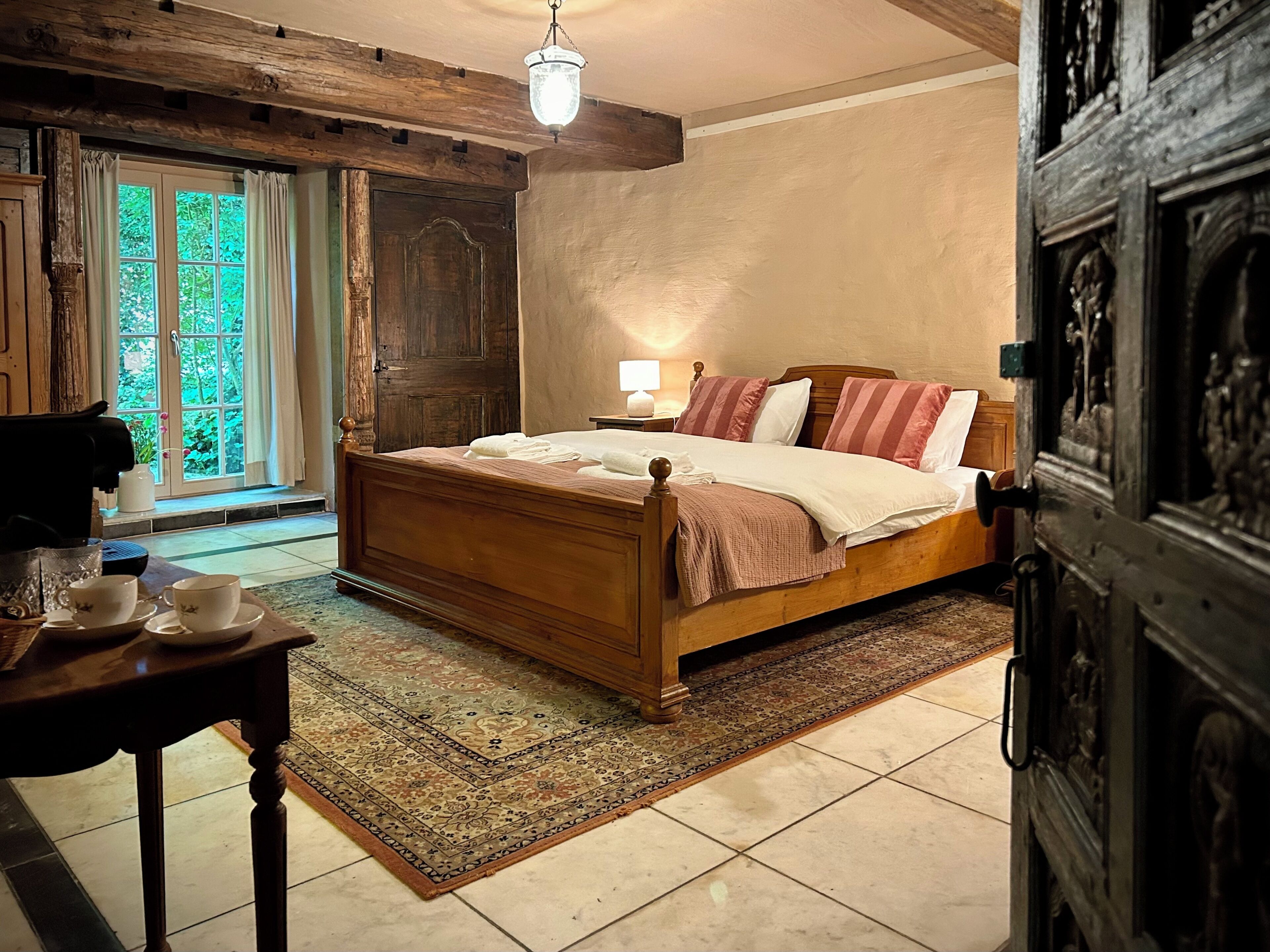 A bedroom with a wooden bed, a nightstand, a table with cups, a lamp, and a view of greenery through a window.