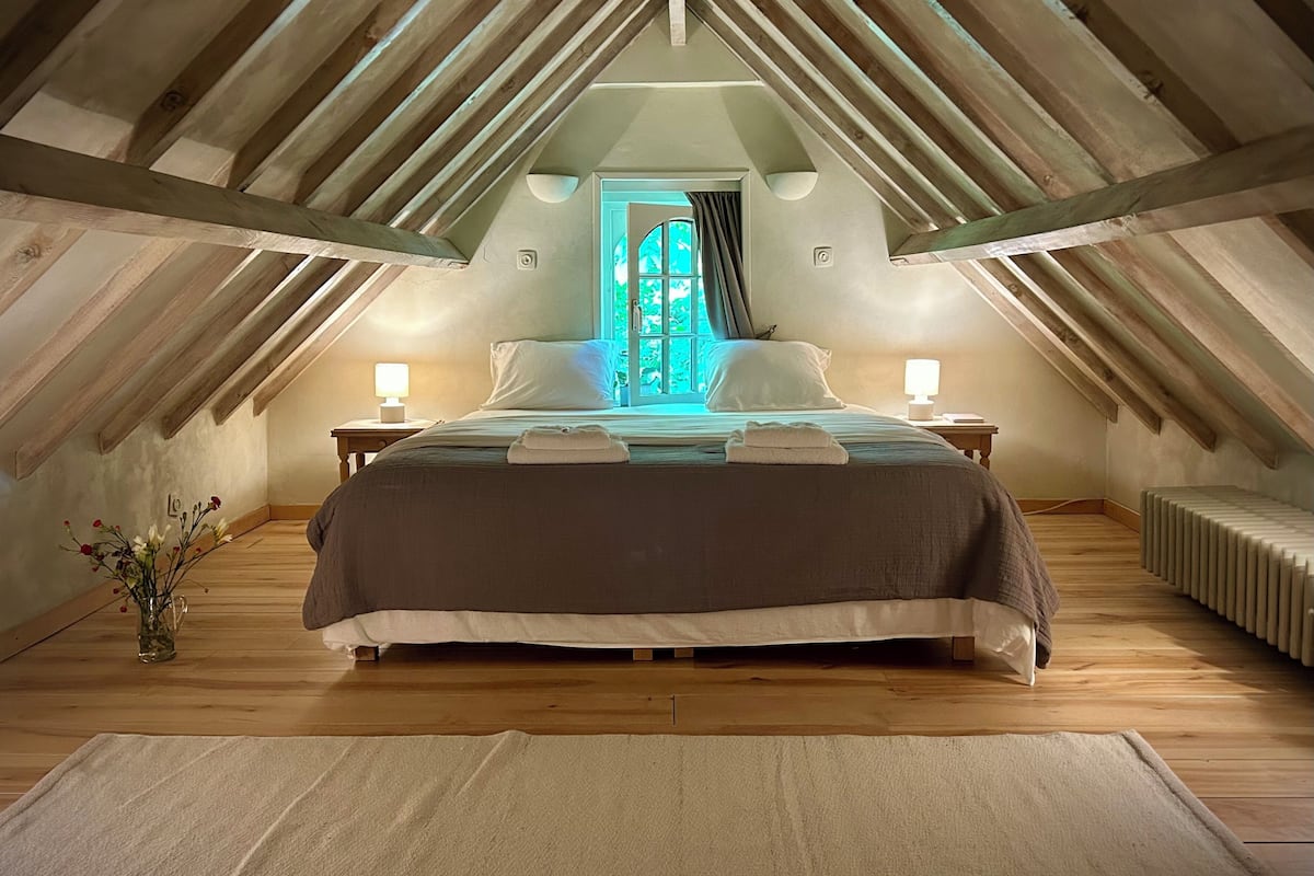 A cozy attic bedroom with a sloped ceiling, a large bed with pillows, bedside lamps, and a view through a window.