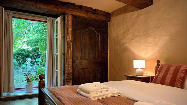 A bedroom with a wooden bed, a window with curtains, a bedside lamp, and a view of greenery outside. - B&B Pronkenburg (Bruges)