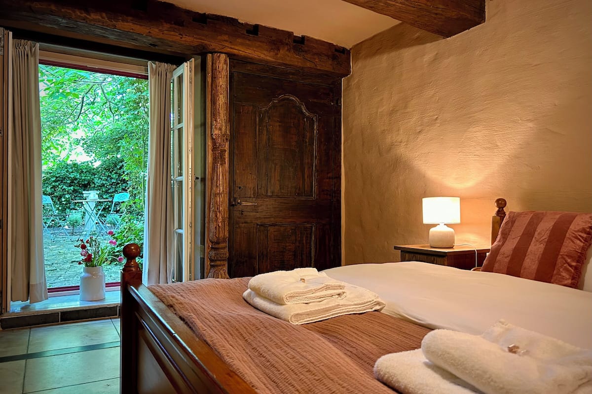 A bedroom with a wooden bed, a window with curtains, a bedside lamp, and a view of greenery outside.