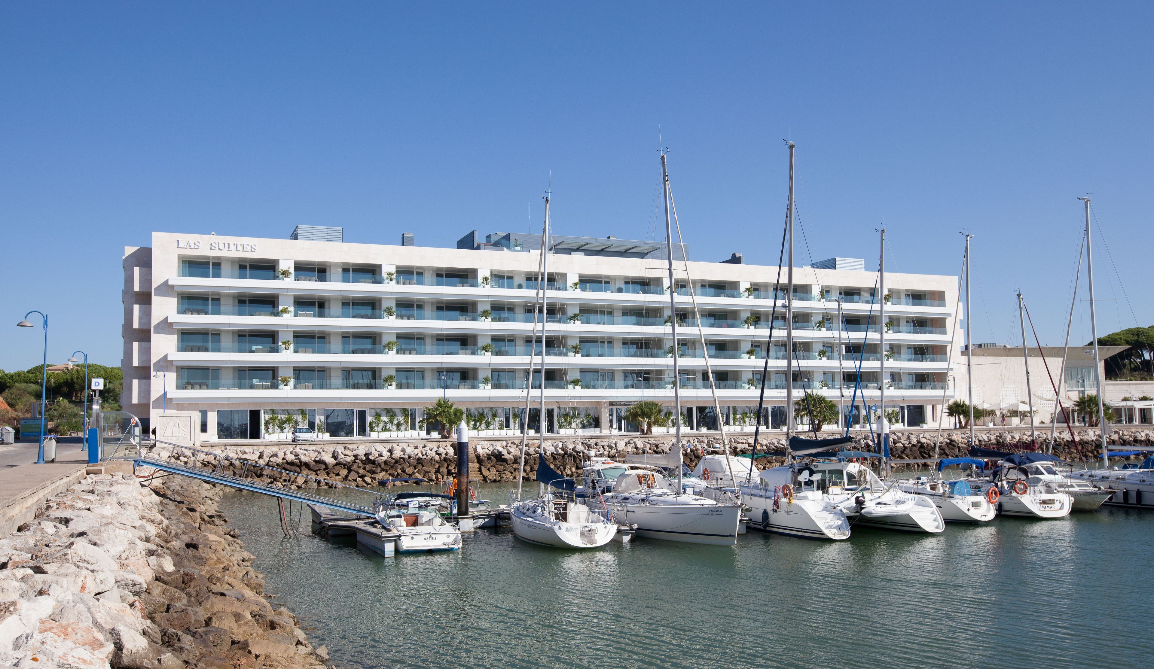 Un hotel de varios pisos con balcones, un puerto deportivo con varios barcos y una costa rocosa.