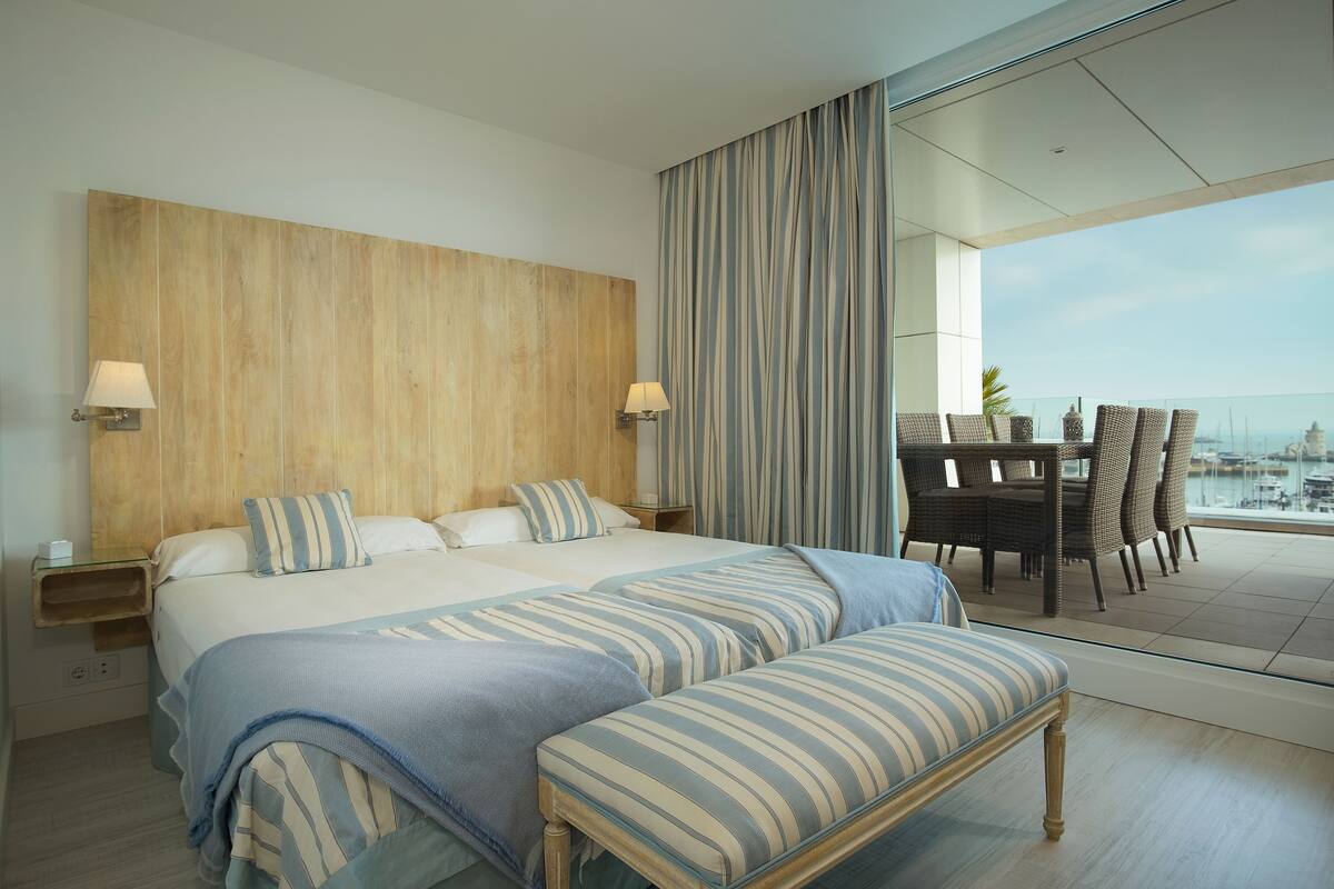 A modern hotel room with a large bed, a bench, a dining area with chairs, and a view of the sea.