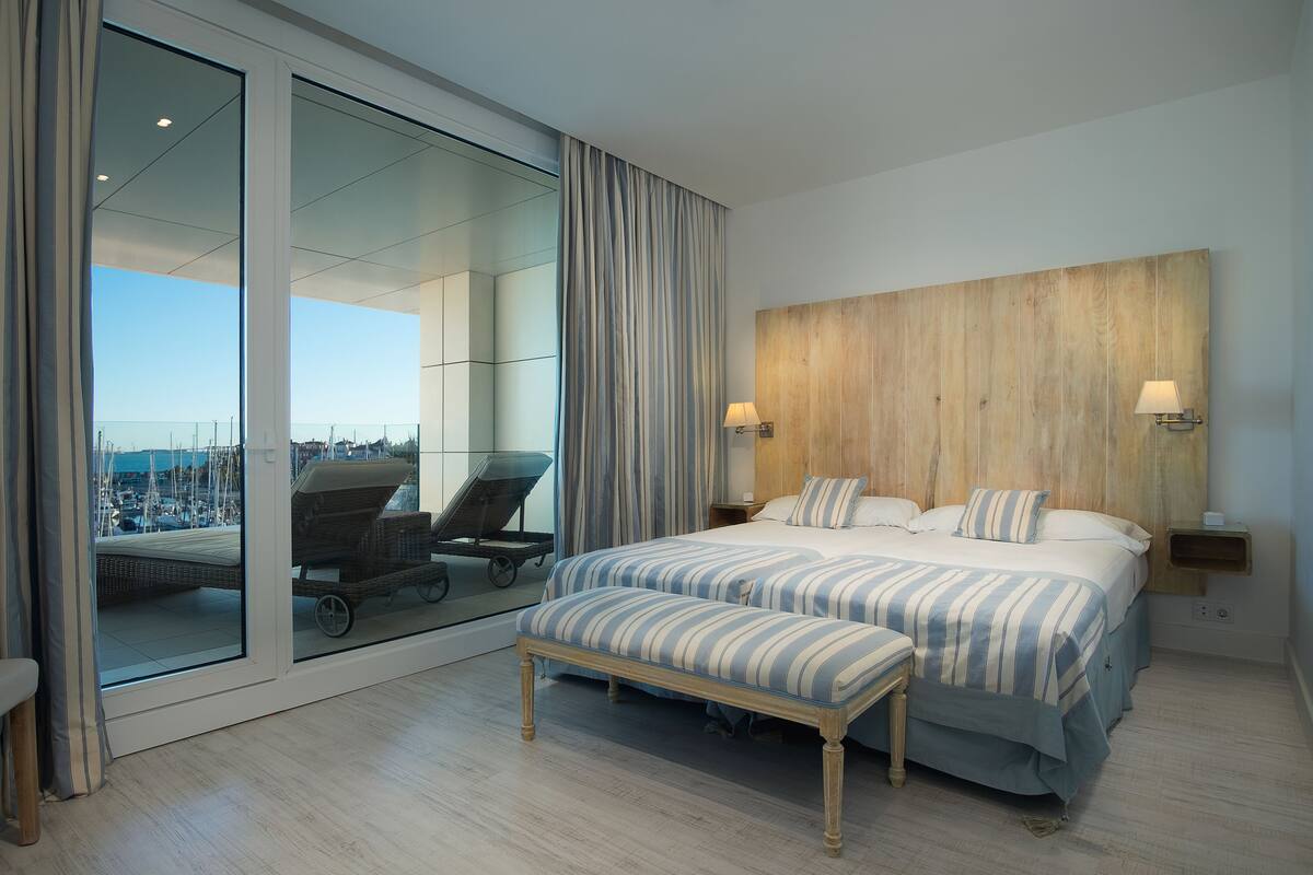 A modern hotel room with a large sliding glass door, a wooden headboard, a bed with striped bedding, a bench, and a view of a marina.