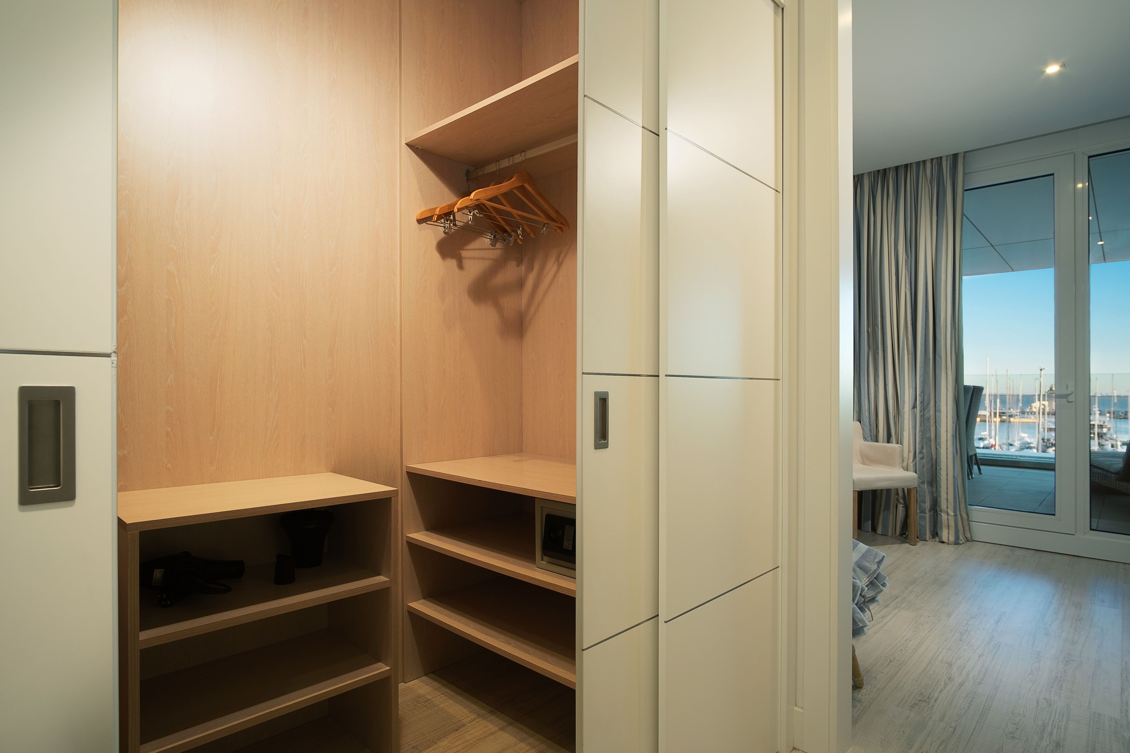 A modern closet with wooden shelves and a hanging rack, a white wardrobe, and a view of a balcony with a cityscape.