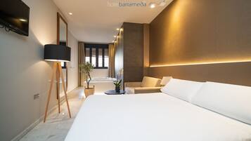 A modern hotel room with a large bed, a sitting area, a small table with a plant, and a bathtub.