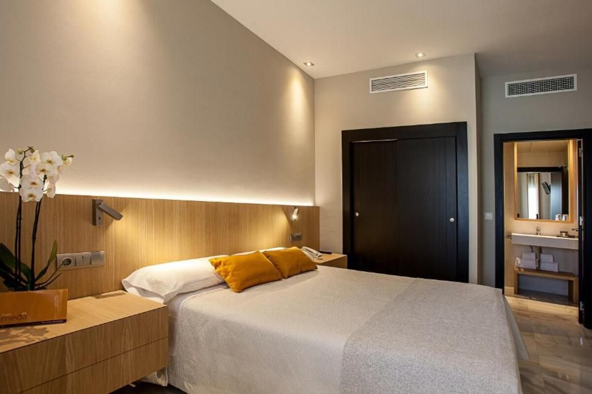 A modern hotel room with a large bed, a bedside table with a lamp, a wooden nightstand, and a mirror above the sink.