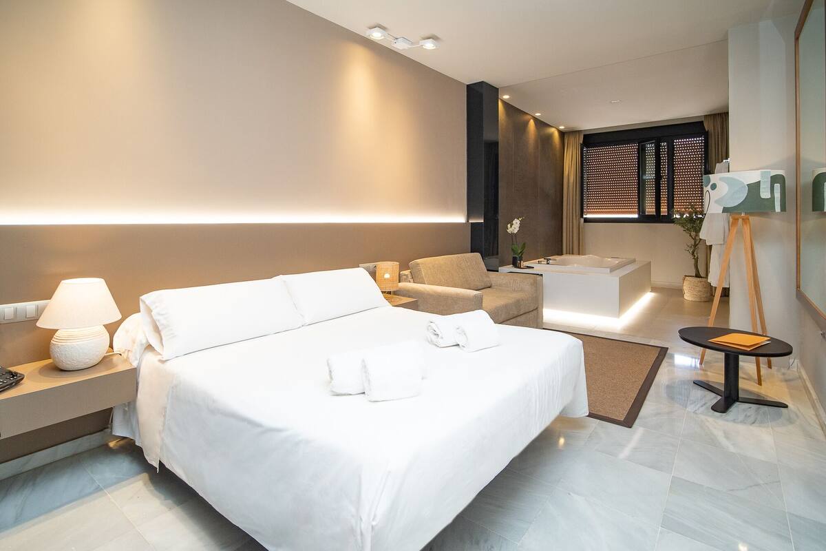 A modern hotel room with a large bed, a sitting area, a bathtub, and a small table with a lamp.