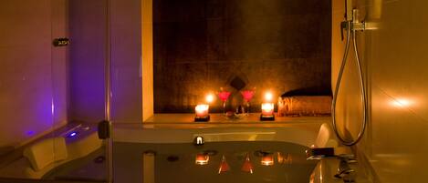 Un baño moderno con bañera de hidromasaje con mampara de vidrio, ducha de mano y velas en el alféizar de la ventana.