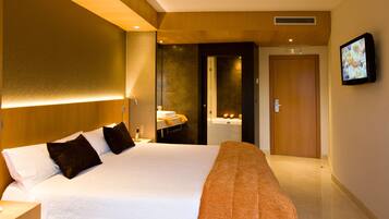 Quarto de hotel com uma cama grande, uma mesinha de cabeceira, uma televisão na parede e um banheiro visível através de uma porta aberta.