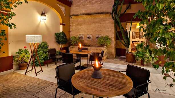 An outdoor patio area with round wooden tables and chairs, potted plants, and a brick wall. - Hotel Barrameda (Sanlucar de Barrameda)