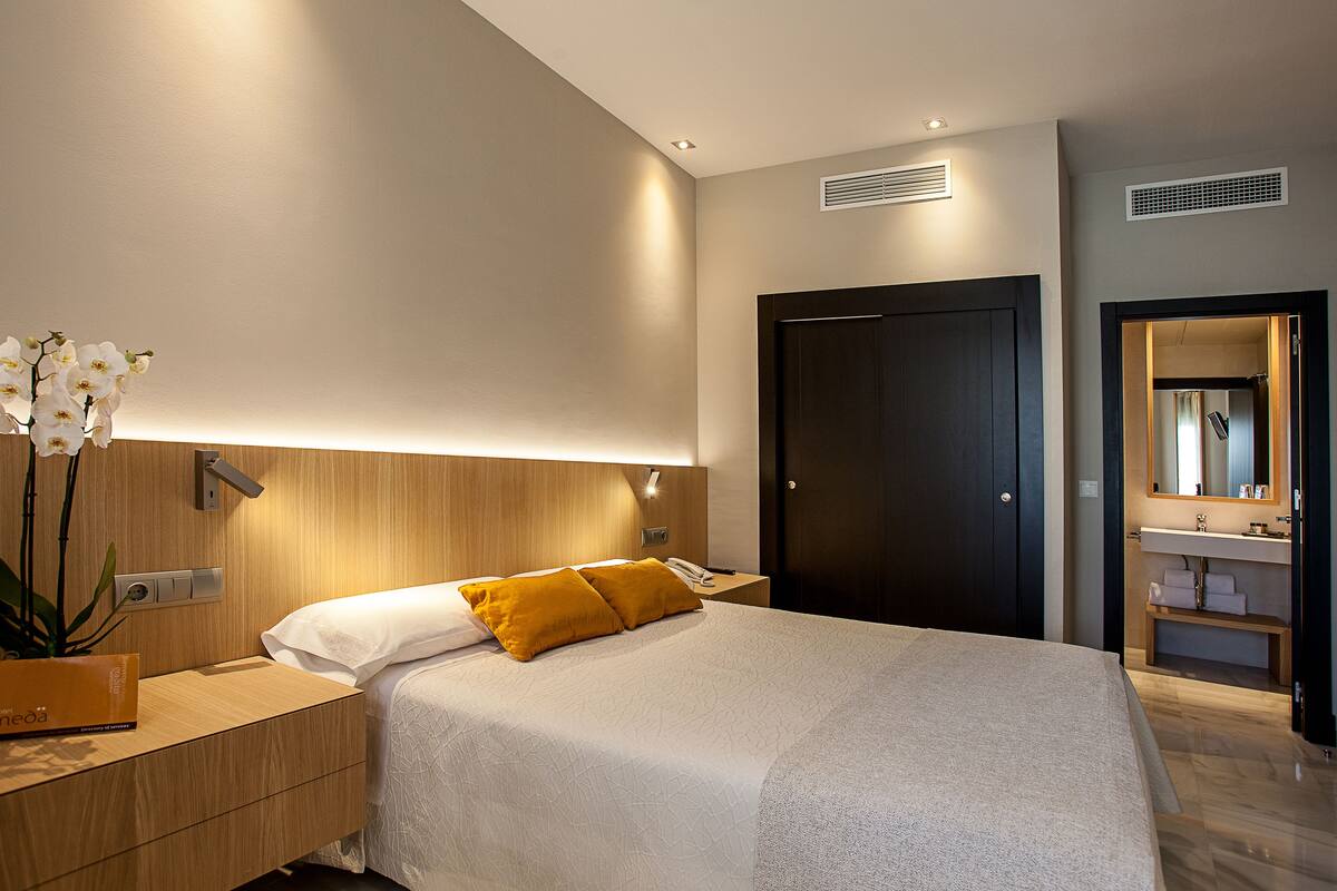 A modern hotel room with a wooden headboard, a bed with white and yellow bedding, a bedside table with a phone, and a potted orchid.