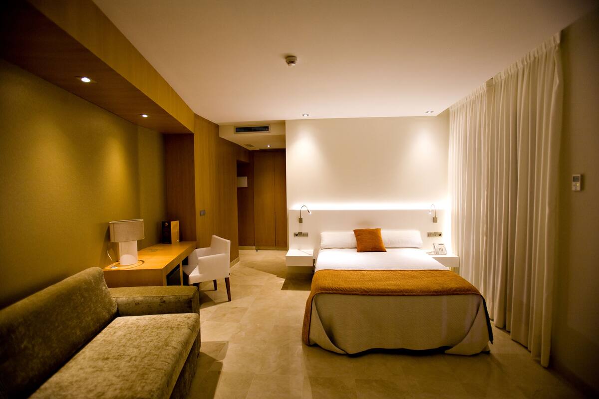A hotel room with a bed, a sofa, a desk with a lamp, and a bedside table.