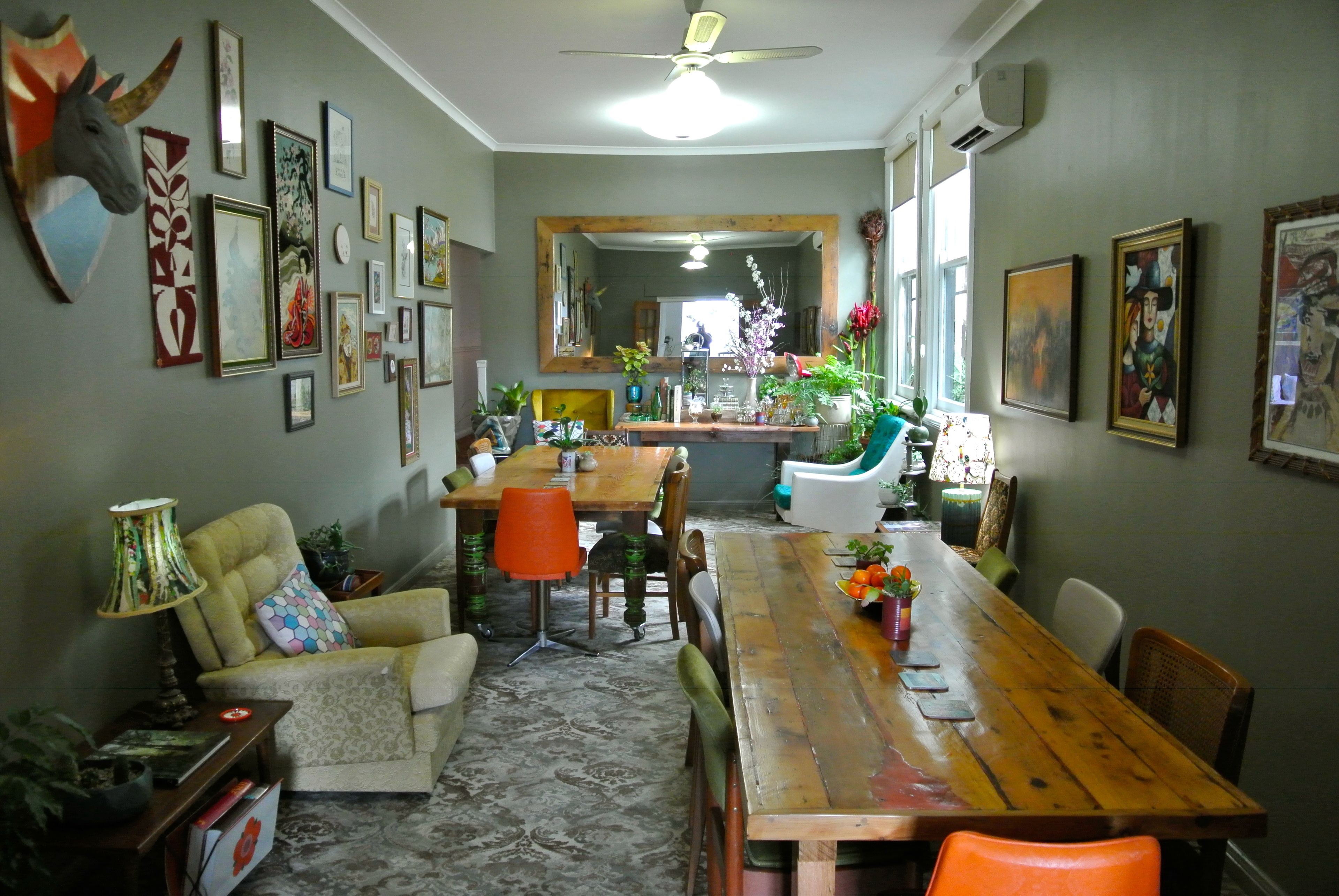 Uma sala de jantar com mesa de madeira, cadeiras e um sofá. Há quadros nas paredes e um ventilador de teto.