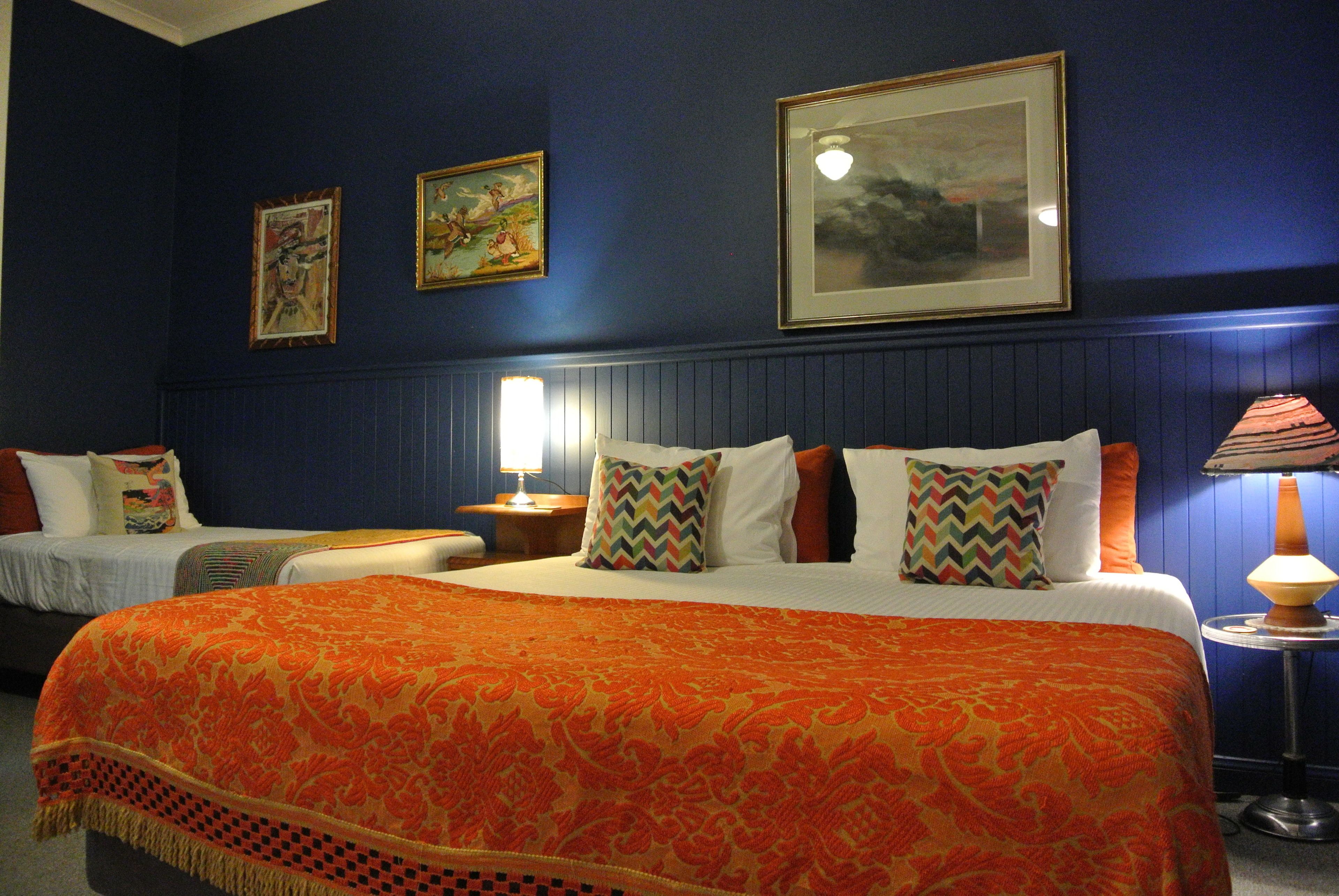 A hotel room with two beds, a large orange patterned bedspread, and a framed landscape painting on the wall.