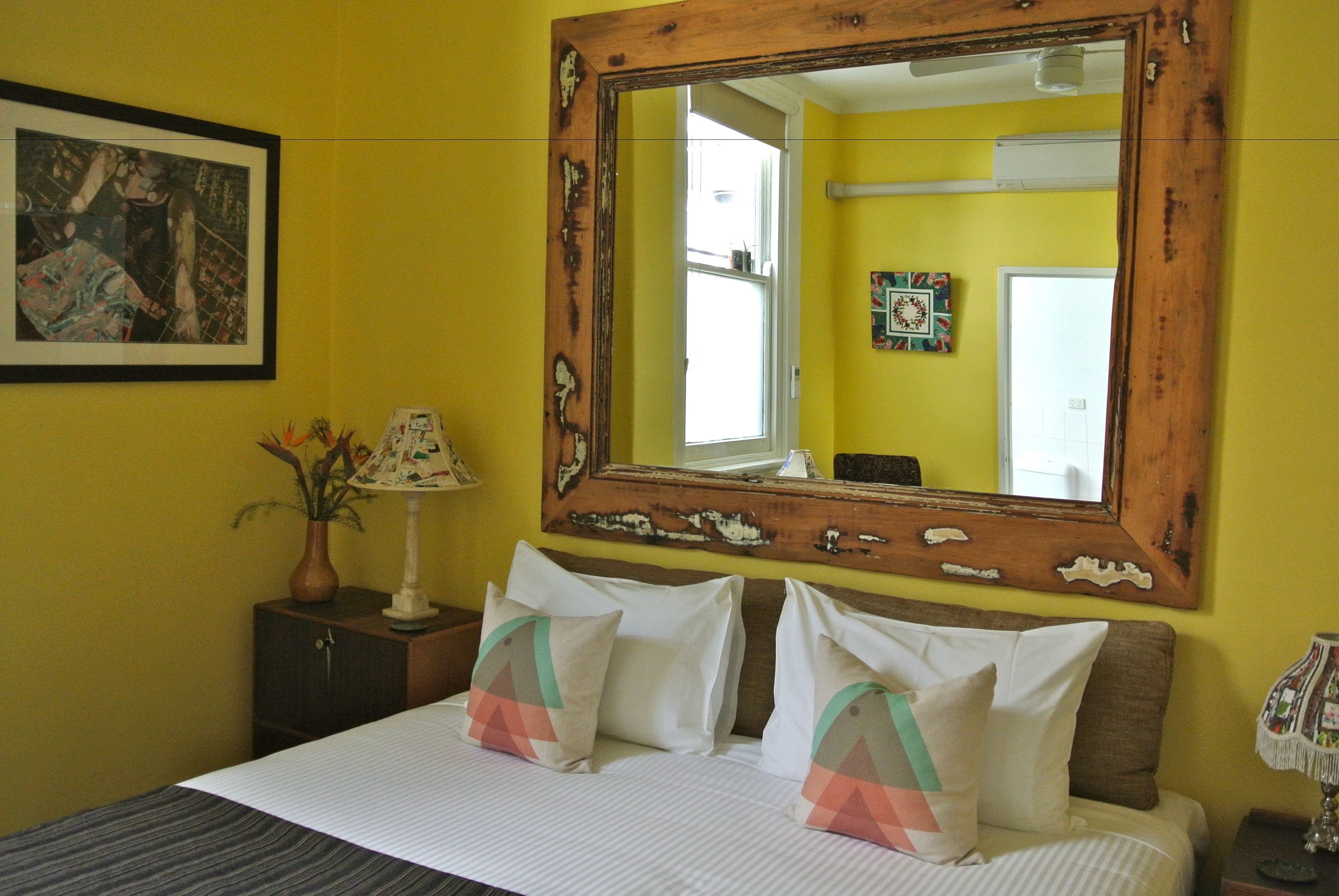 A bedroom with a large mirror, a bed with patterned pillows, a nightstand with a lamp, and a framed picture on the wall.