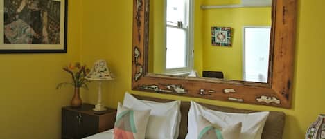 A bedroom with a large mirror, a bed with patterned pillows, a nightstand with a lamp, and a framed picture on the wall.