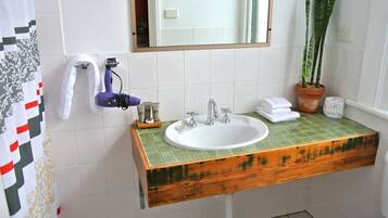 Un baño con lavamanos blanco, tocador de madera, encimera con azulejos verdes, una planta en maceta, un espejo y cortina de ducha.