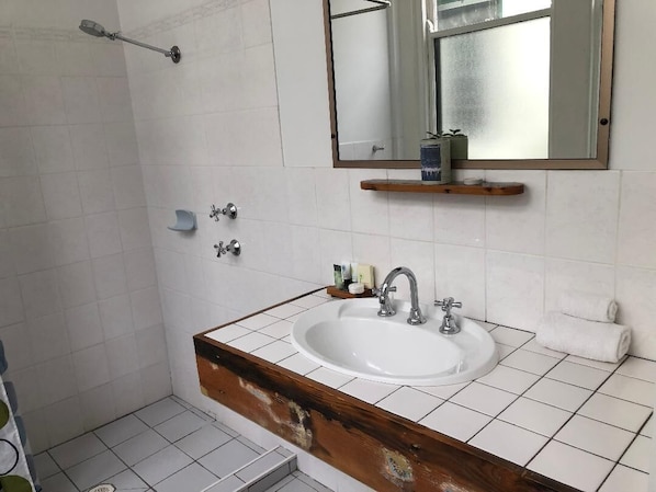 Un baño con lavamanos blanco, tocador de madera, espejo y zona de ducha.