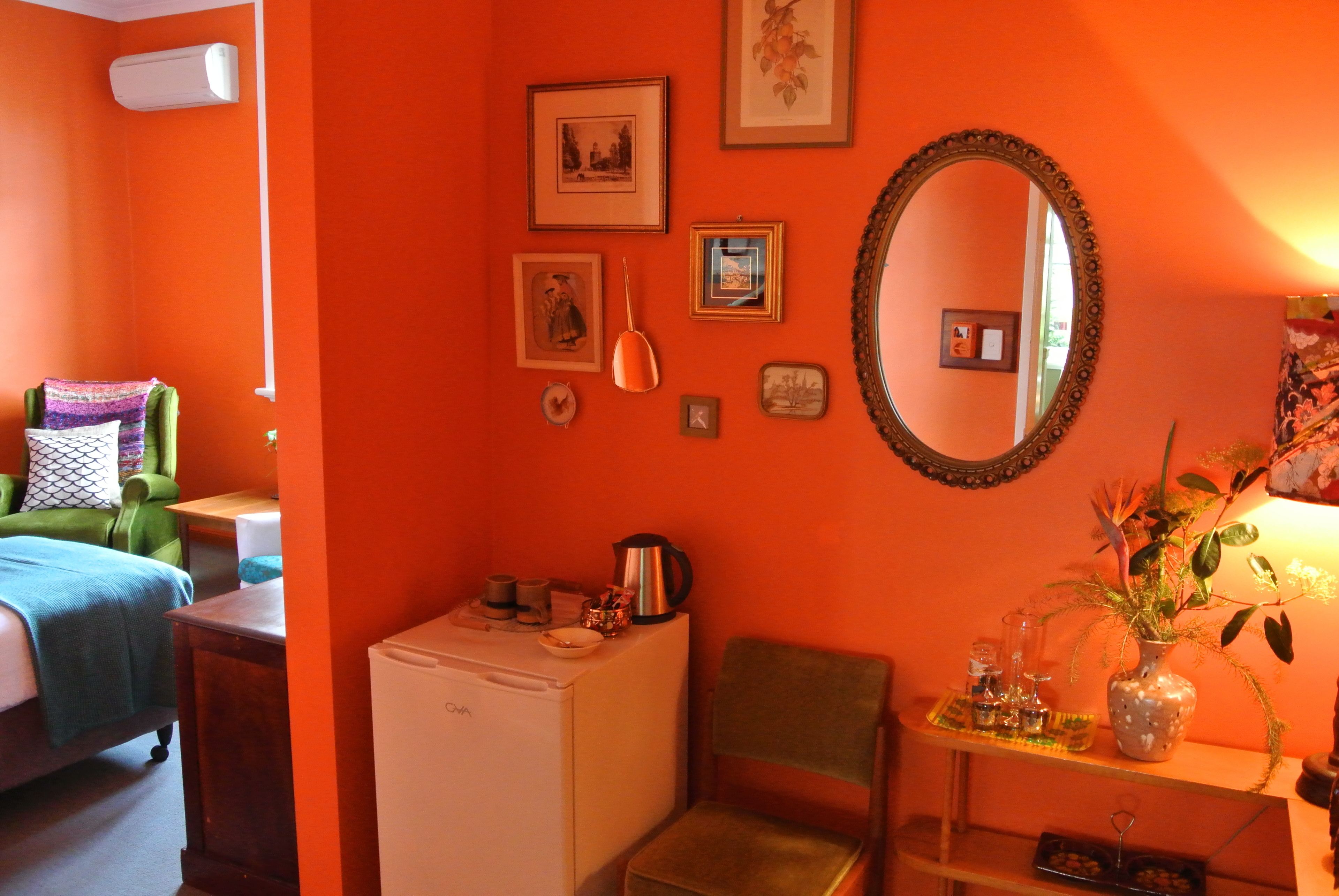 Una habitación con cama, un refrigerador pequeño, una silla, una mesa con un florero y un espejo en la pared.