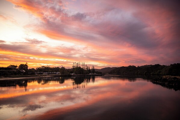 A calm lake reflecting a sunset sky with trees and buildings in the background. - Reflections Massy Greene - Holiday Park (Brunswick Heads)