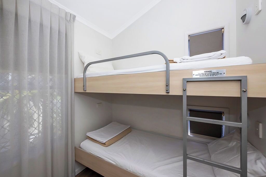 Un lit superposé avec un ordinateur portable posé sur le lit du haut, une petite fenêtre et un panneau d’avertissement indiquant la hauteur du lit.