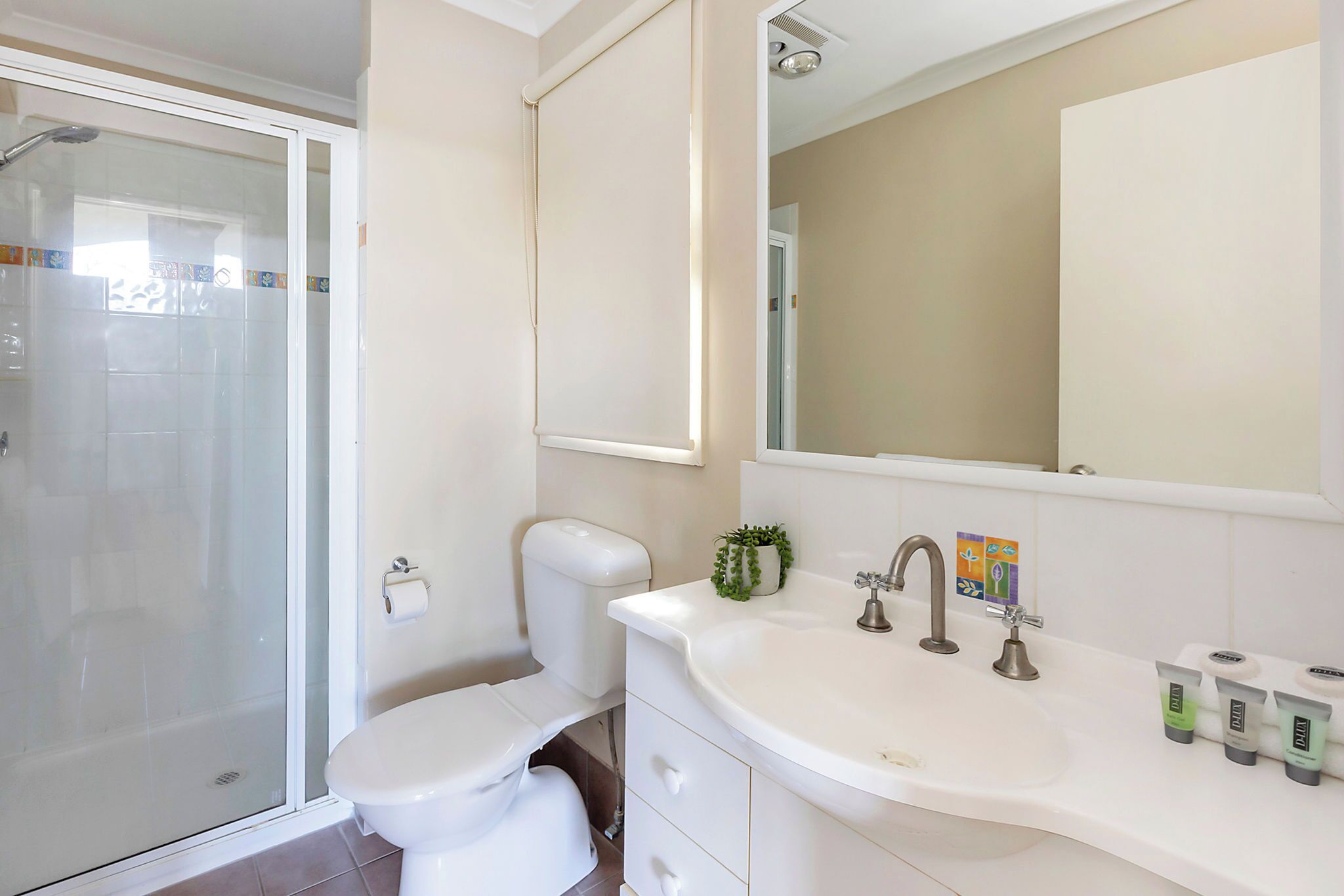 A bathroom with a glass shower enclosure, a toilet, a sink with a mirror, and toiletries on the sink.