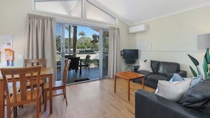 A spacious living area with a sliding glass door leading to a balcony, a black leather sofa, a wooden dining table with chairs, and a flat-screen TV mounted on the wall. - Reflections Brunswick Head - Holiday Park (Brunswick Heads)