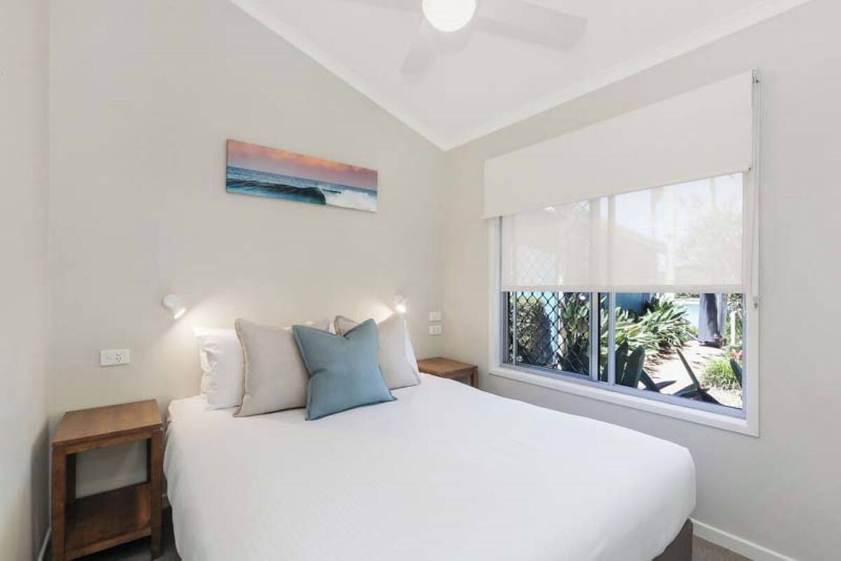 Ein Schlafzimmer mit einem großen Bett, einem Nachttisch, einer Wandlampe, einem Deckenventilator und einem Fenster mit Blick ins Freie.