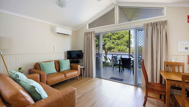 A living room with a brown leather sofa, a television, and a wooden dining table with chairs. - Reflections Brunswick Head - Holiday Park (Brunswick Heads)