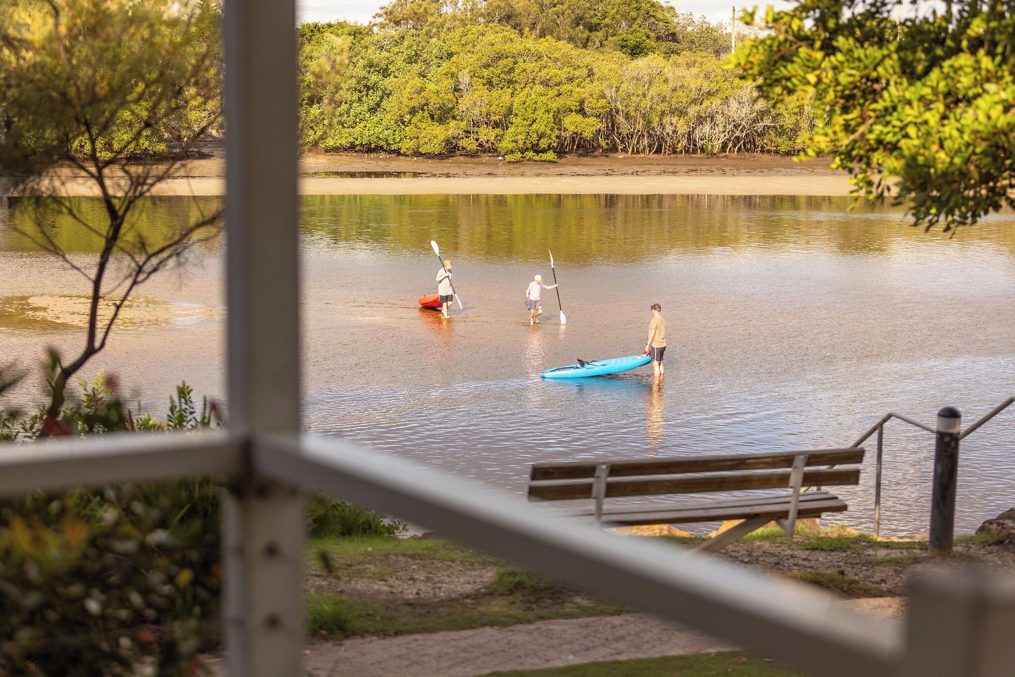 Un lago tranquilo con gente remando en kayak, un banco de madera y una barandilla.