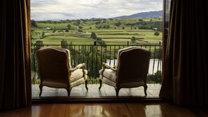 A room with two upholstered chairs facing a balcony with a scenic view of a green landscape and a body of water. - Lake Karapiro Lodge (Karapiro)