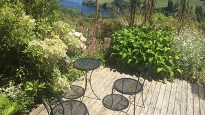 A wooden deck with two round metal tables and chairs, surrounded by lush greenery and a scenic view of a lake and hills. - Lake Karapiro Lodge (Karapiro)