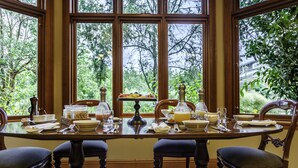 A dining table set for four with a view of a lush garden through large windows. - Lake Karapiro Lodge (Karapiro)