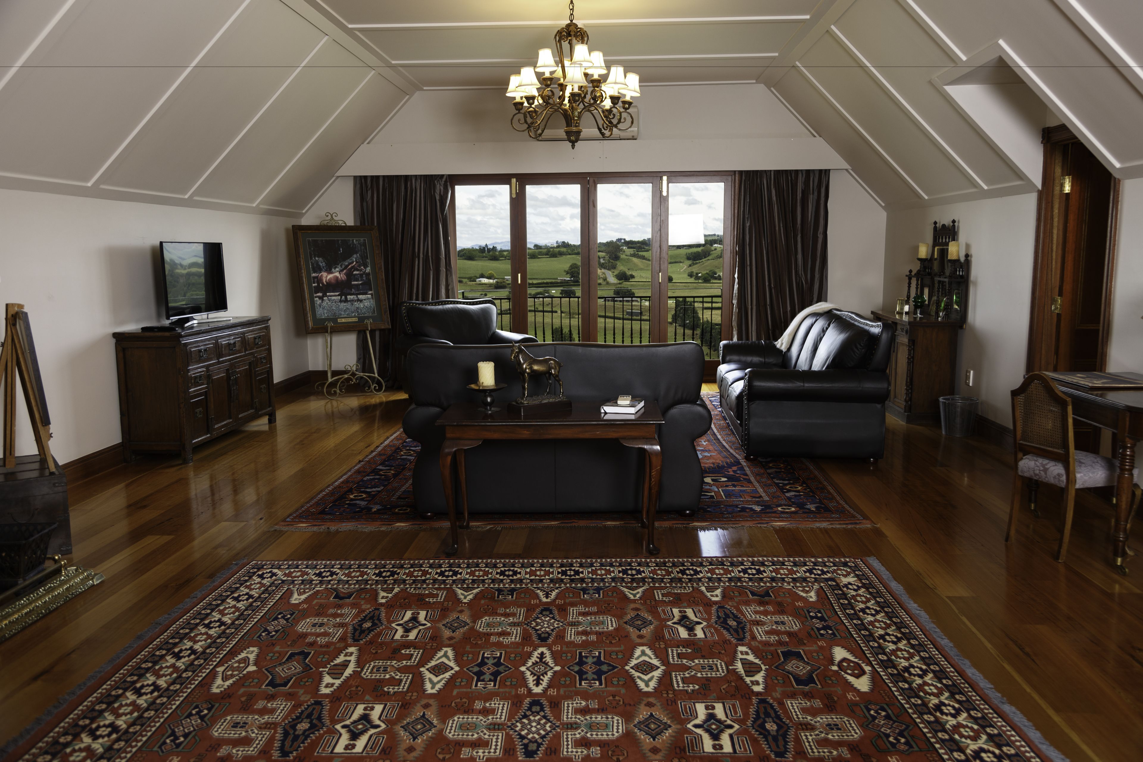 A spacious living room with a large window, a chandelier, and a traditional rug.