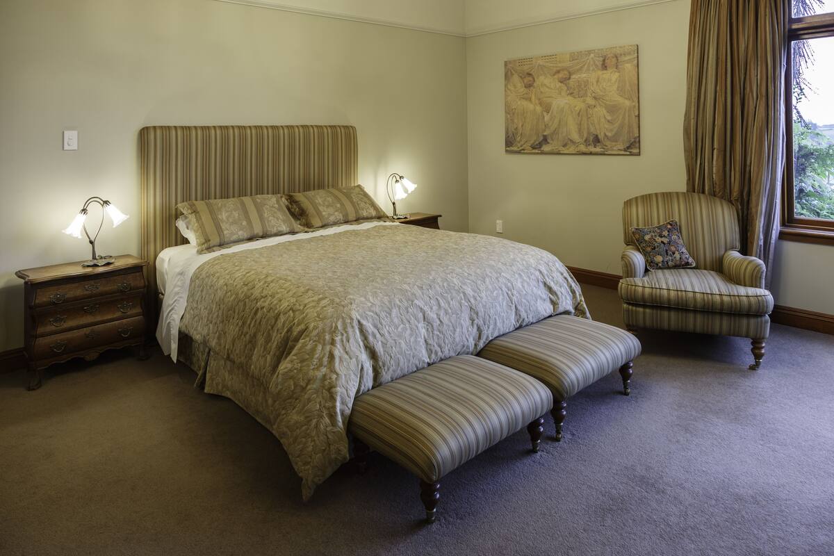 A bedroom with a large bed, a nightstand with a lamp, a chair with a cushion, and a painting on the wall.