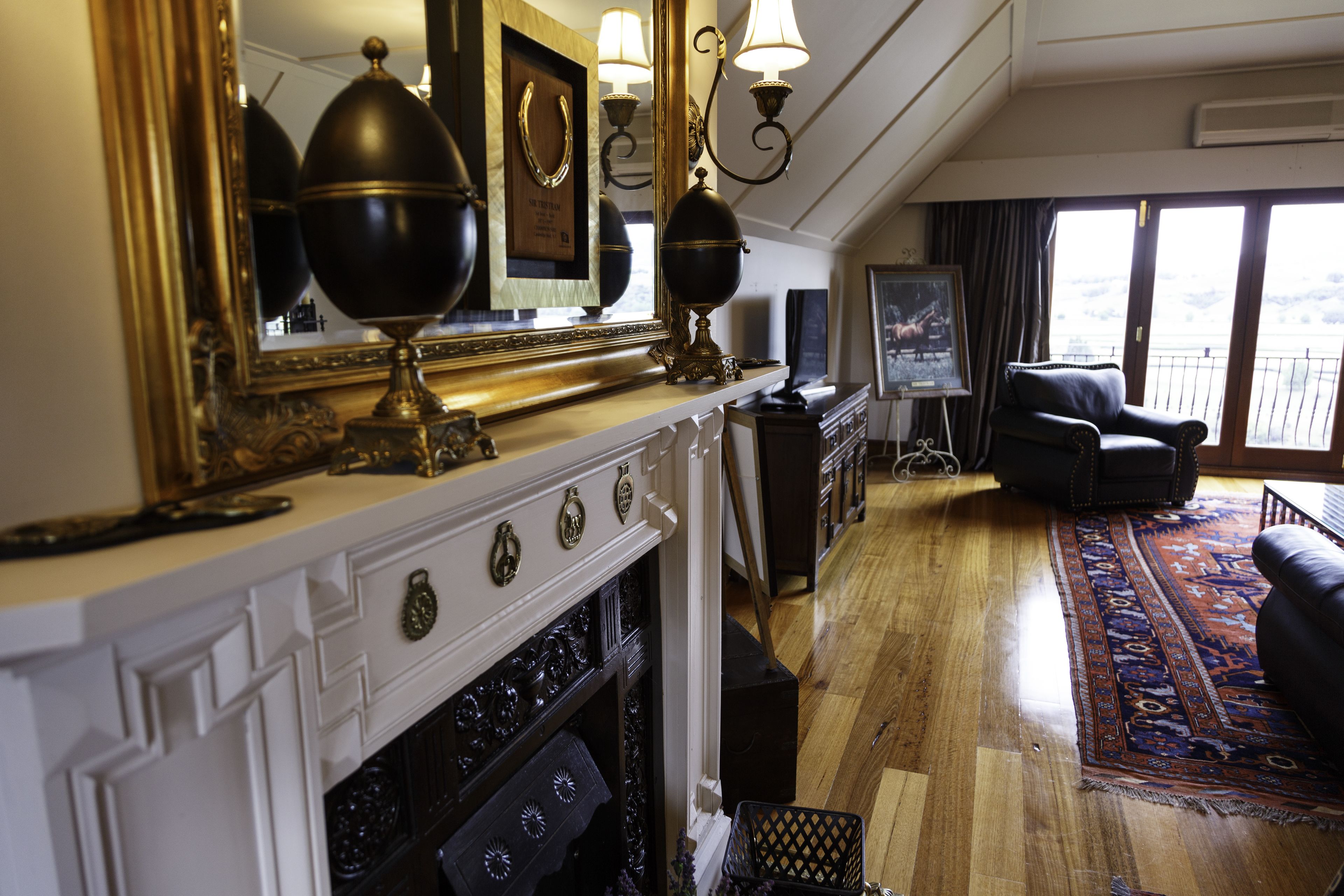 A room with a fireplace, a large mirror, a black leather sofa, and a view of the outdoors through a large window.