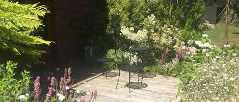 A wooden deck with two round tables and chairs, surrounded by lush greenery and flowering plants.