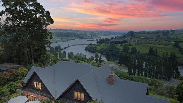 A house with a dark roof, large windows, and a view of a river and hills. - Lake Karapiro Lodge (Karapiro)