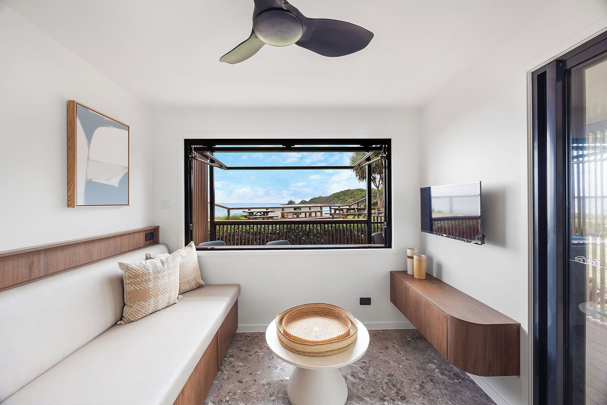 Une chambre moderne avec un canapé, une petite table sur laquelle se trouve un panier, un ventilateur de plafond, une fenêtre donnant sur un balcon et un téléviseur fixé au mur.