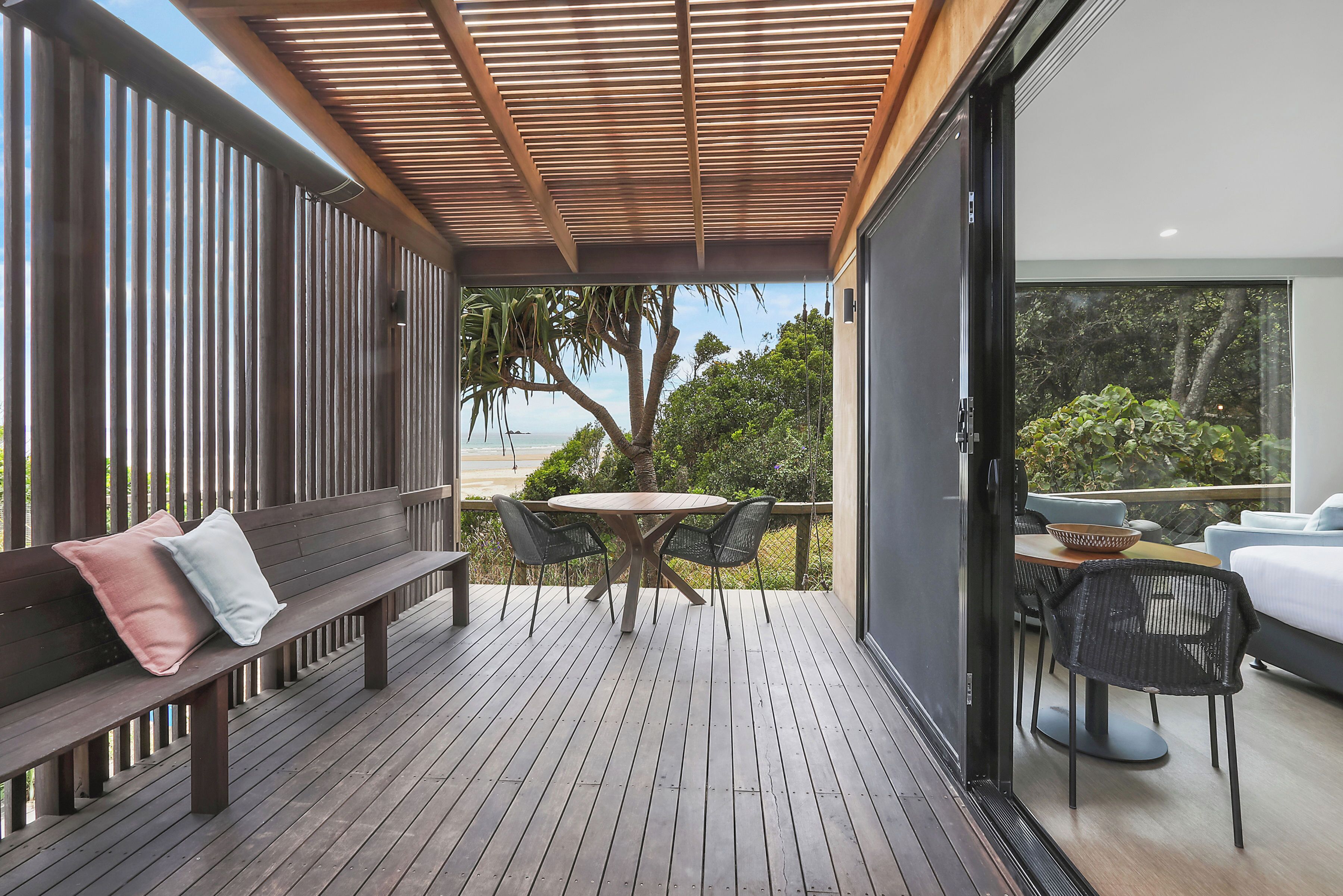 Eine Holzterrasse mit einem runden Tisch und Stühlen, einer Bank mit Kissen sowie Blick auf Grünflächen und ein Gebäude.