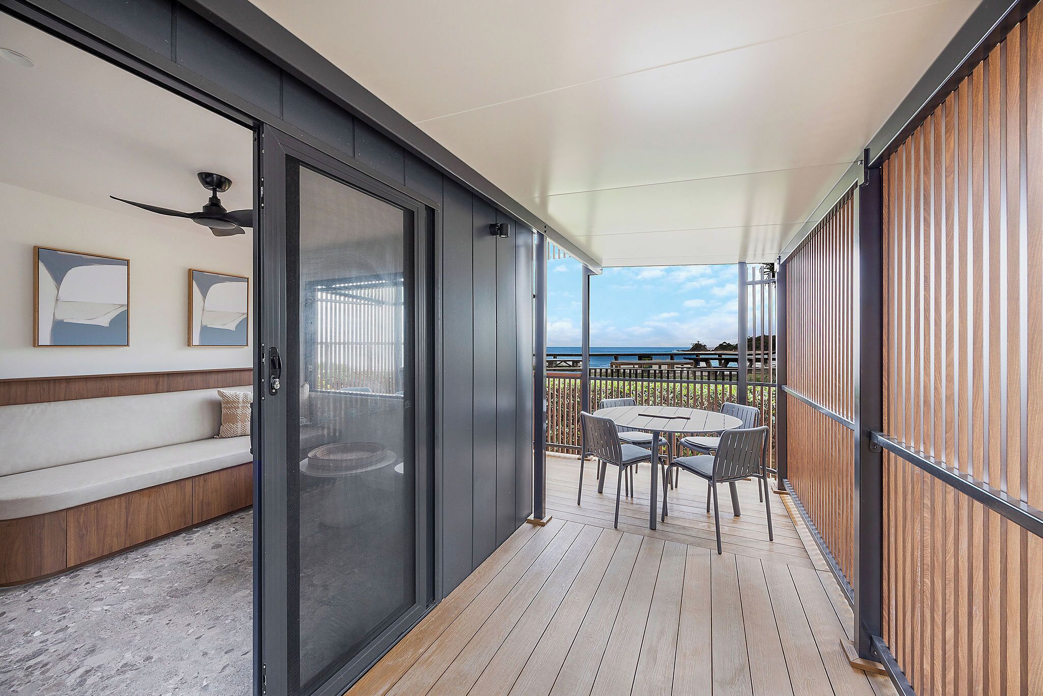 A covered outdoor area with a wooden deck, a seating area with a sofa and cushions, a table with chairs, and a view of the sea.