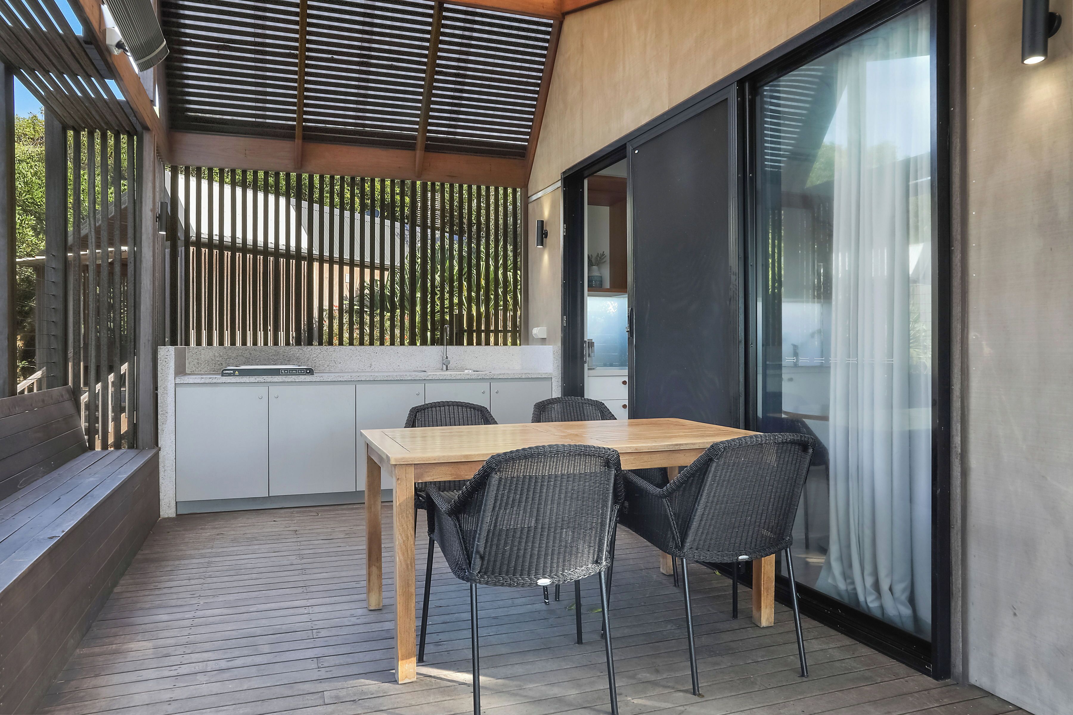 Een moderne buiten eethoek met een houten tafel en zwarte rieten stoelen, een schuifdeur en uitzicht op een tuin.