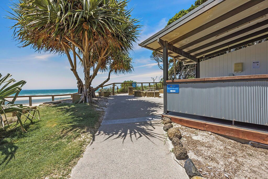 A pathway leading to a beach access point, with a palm tree and a building with a sign.