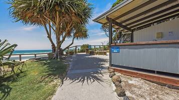 A pathway leading to a beach access point, with a palm tree and a building with a sign.